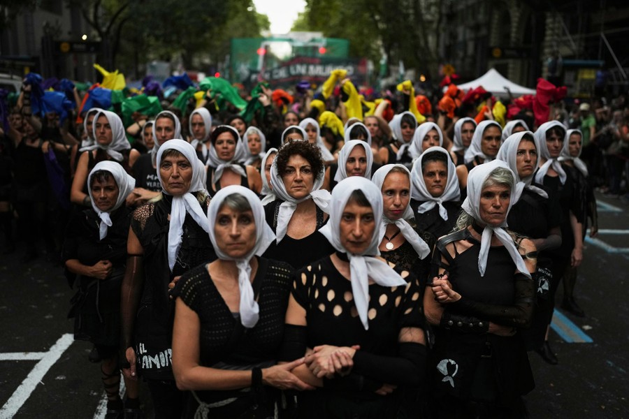 Women wearing white scarves and black clothing lock arms together while walking down a street during a commemoration ceremony.