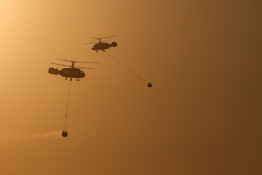 Two firefighting helicopters pass each other carrying buckets attached to long cables.