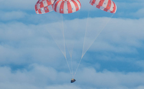 A gumdrop-shaped spacecraft, charred from the heat of reentry, coasts through blue sky beneath a trio of red-and-white parachutes.