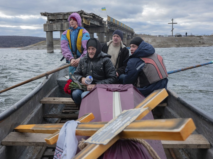 Four civilians sit in a small boat alongside a coffin, crossing a river near a destroyed road bridge.