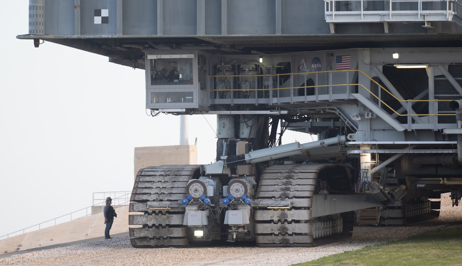 A person stands on a gravel road beside one of several gigantic tracks that belong to a large vehicle that towers above him.