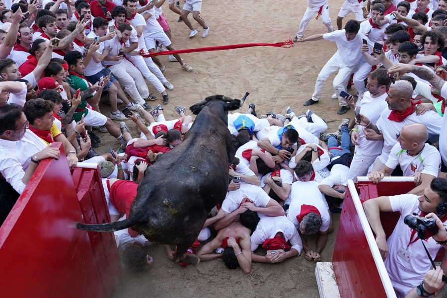 A group of people lies in a small pile at the entrance to a bullring, as a running steer leaps over them.