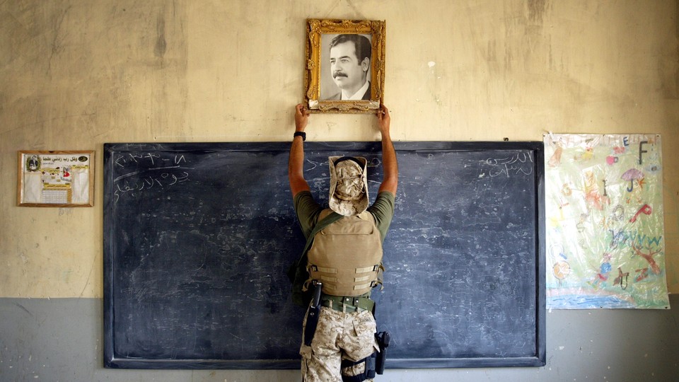 A U.S. Marine, photographed from behind, raises his hands to grip the frame of a portrait of Saddam Hussein hanging above a blackboard.