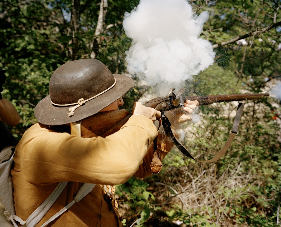 photo of a reenactor wearing a leather brimmed hat with rope tie and tan jacket kneeling and firing a musket through trees, with a cloud of smoke