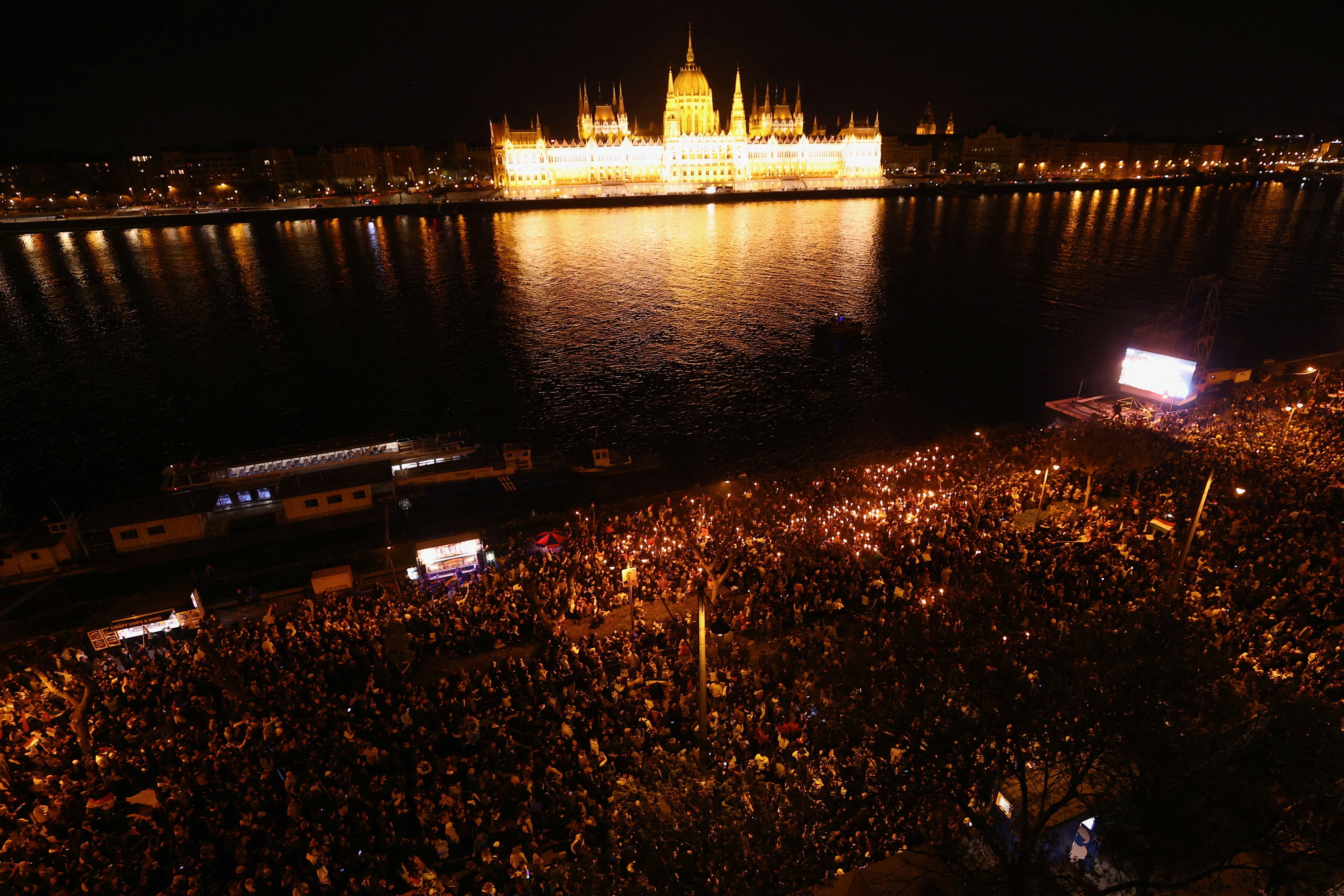 An aerial view of a crowd gathered on the Danube River, with Hungary's parliament building lit up on the opposite side of the river.