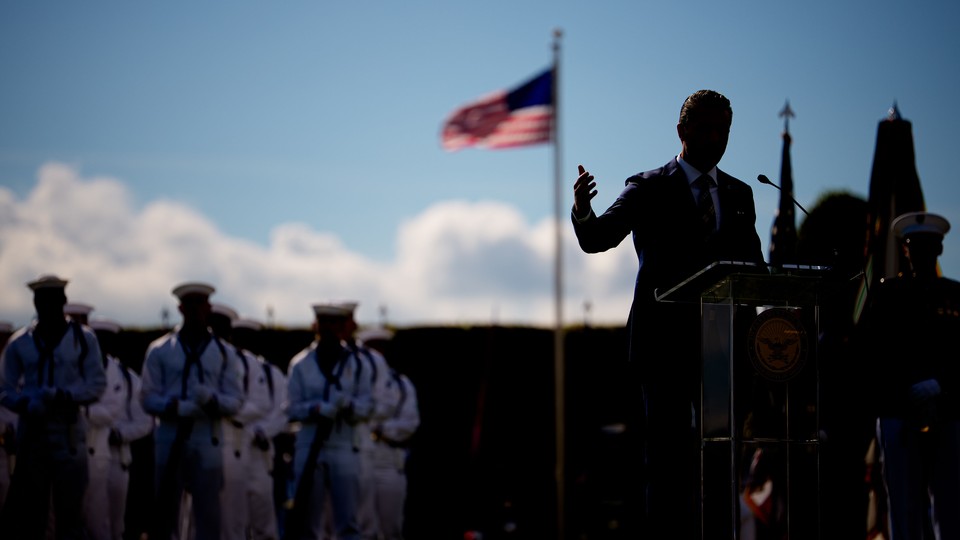 A darkly lit photo of Hegseth speaking at a podium, with an American flag and military cadets lined up behind him