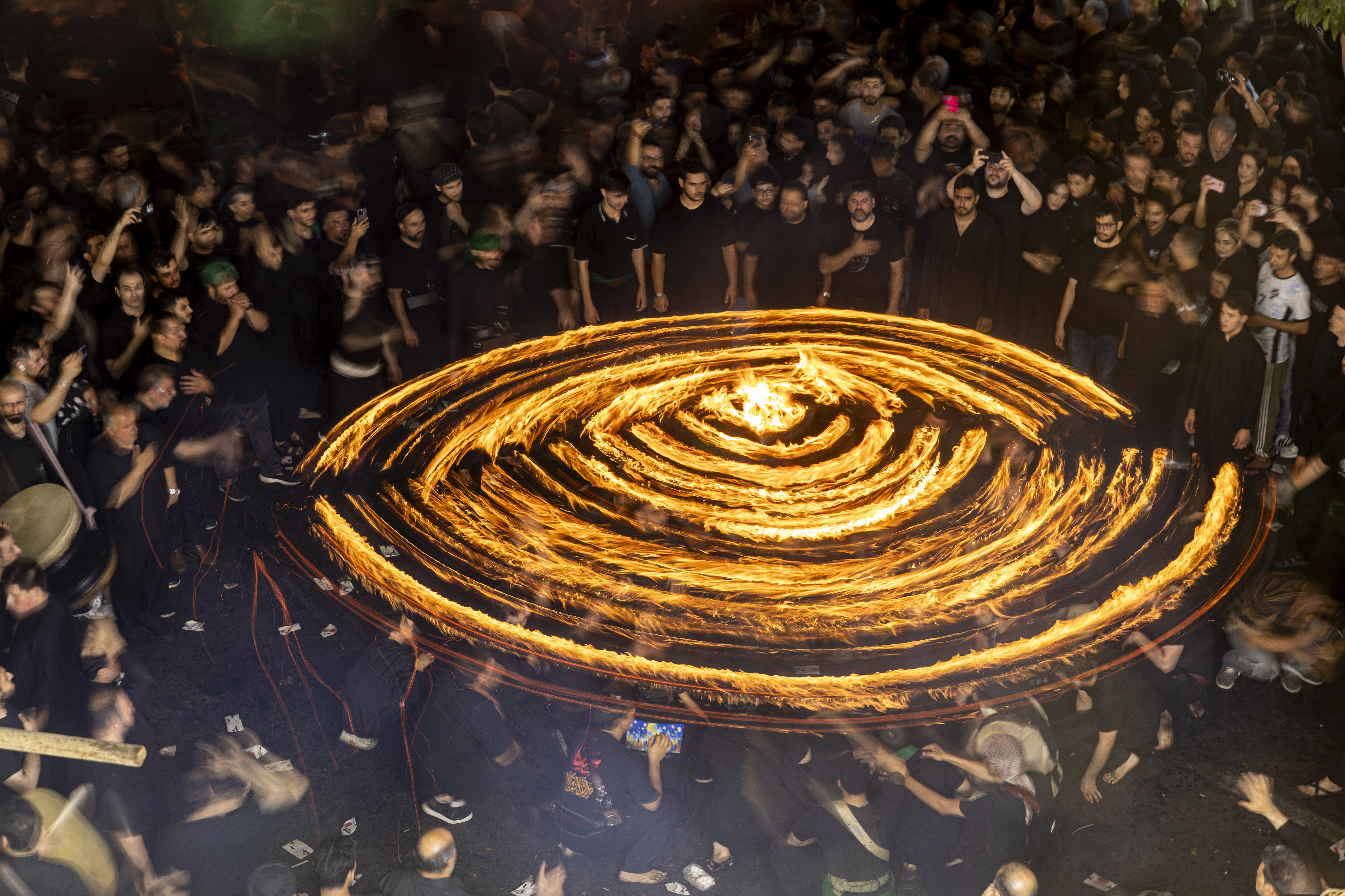 A group of men gather around others who spin a long board with flaming torches attached.