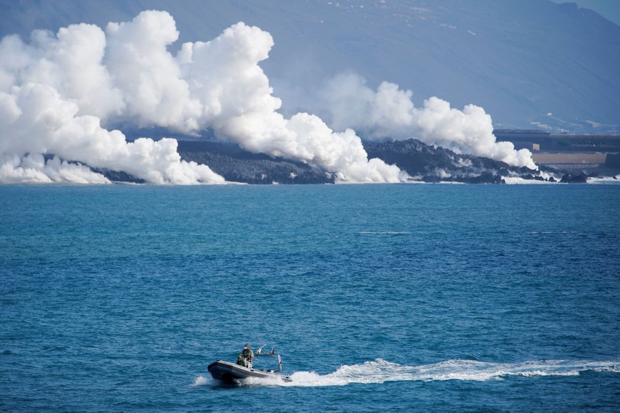 A view across a bay, of a lava flow reaching the seashore, sending clouds of steam into the air.