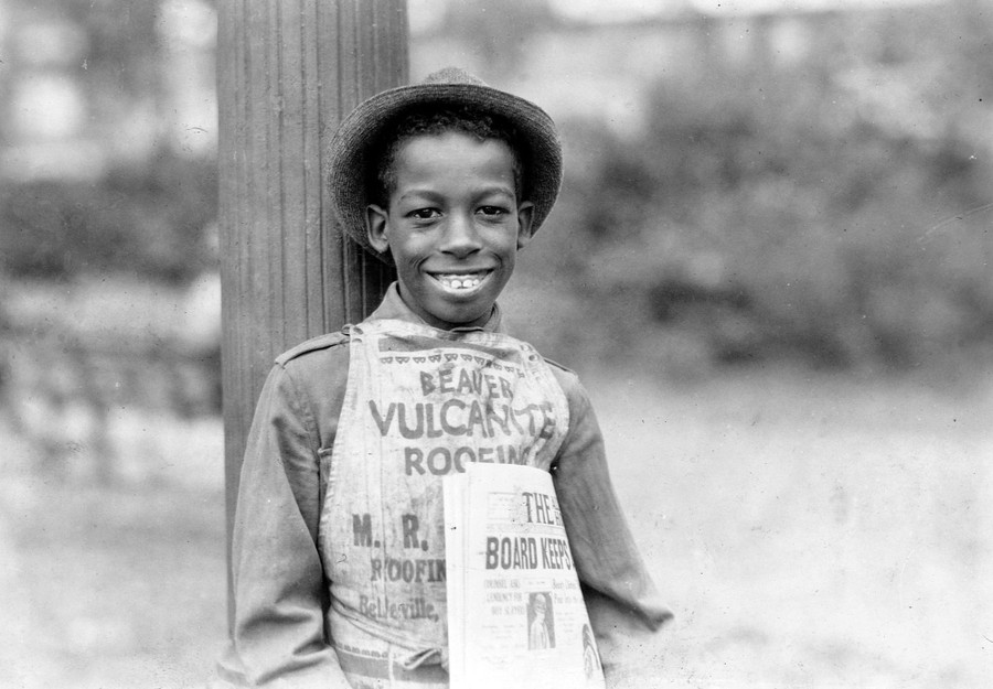 A young newspaper carrier wearing a hat and apron leans against a post while holding a newspaper.