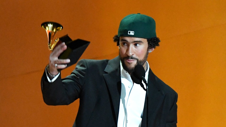 Bad Bunny, wearing a white button-up shirt, a dark suit jacket, and a green baseball cap turned backwards, stands in front of a microphone holding a Grammy award.