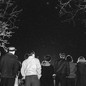 A black-and-white photograph of a group of people looking up at the night sky, which is framed by some trees