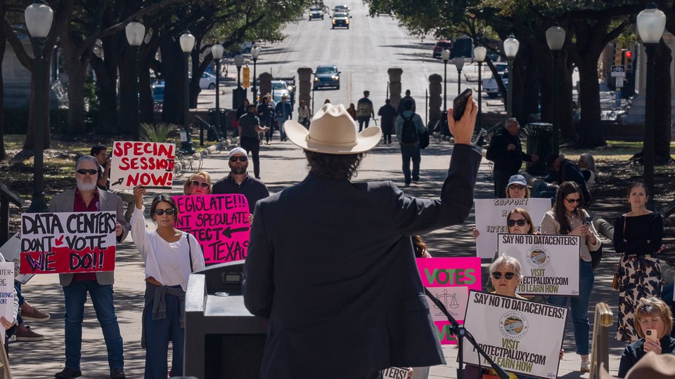 A man in a cowboy hat stands with his phone raised in front of anti-AI protesters
