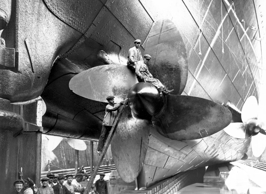 Two workers sit atop one of the huge propellers of a large ship in dry dock.