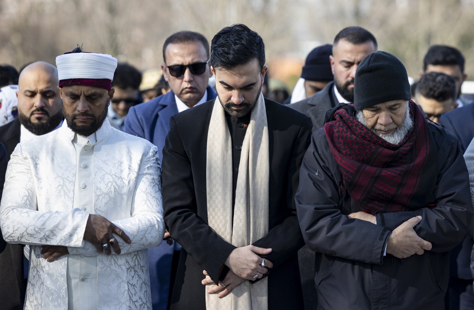 A group of men stand shoulder to shoulder, praying in a park, with New York City Mayor Zohran Mamdani standing at center.
