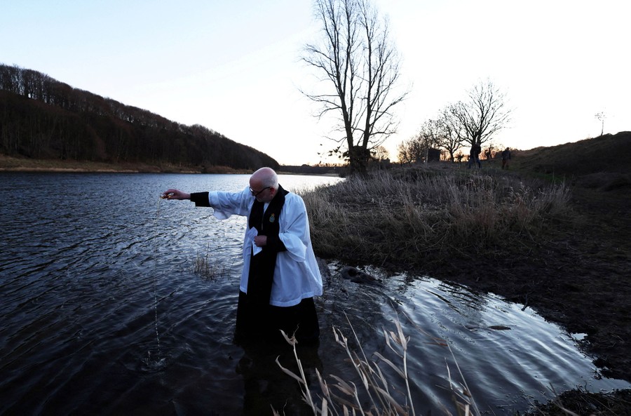 A reverend stands knee-deep in a small body of water, pouring a vial of water into it.