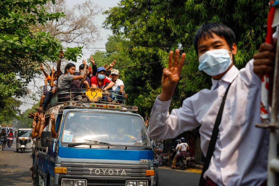 People raise their hands in a salute as they ride on overcrowded vans in a city street.
