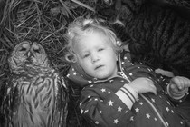 A black-and-white photo of an owl and a child lying in the grass, staring into the lens of the camera.