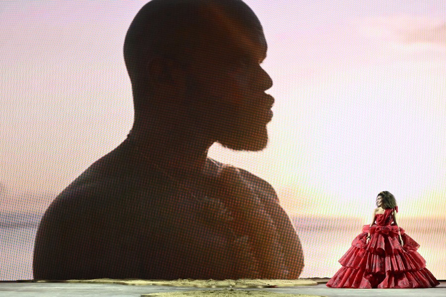 A model walks in a voluminous red dress in front of a large screen displaying a picture of a man.