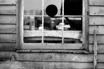 A black and white photo of a window in a wooden house, showing a table inside with jars on it
