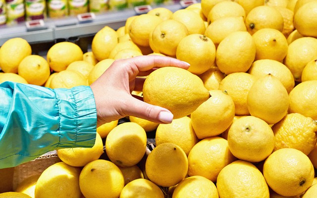 A color photo of someone picking up a lemon from a pile of them in a grocery store.