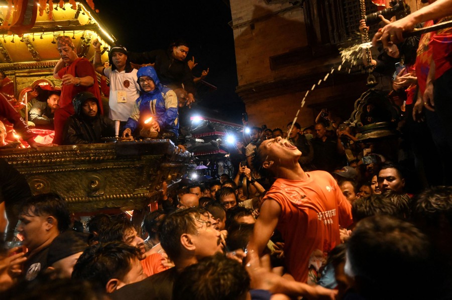 A person stretches up, their mouth wide, to drink wine being poured from above, among a crowd of revelers at a festival.