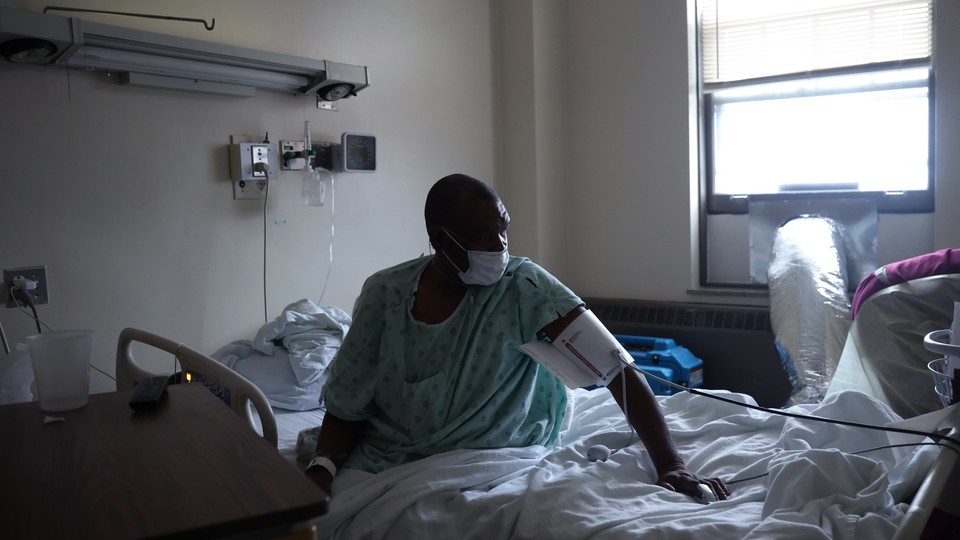 A masked man in a green gown sits up in a hospital bed, wearing medical cuff around his upper arm