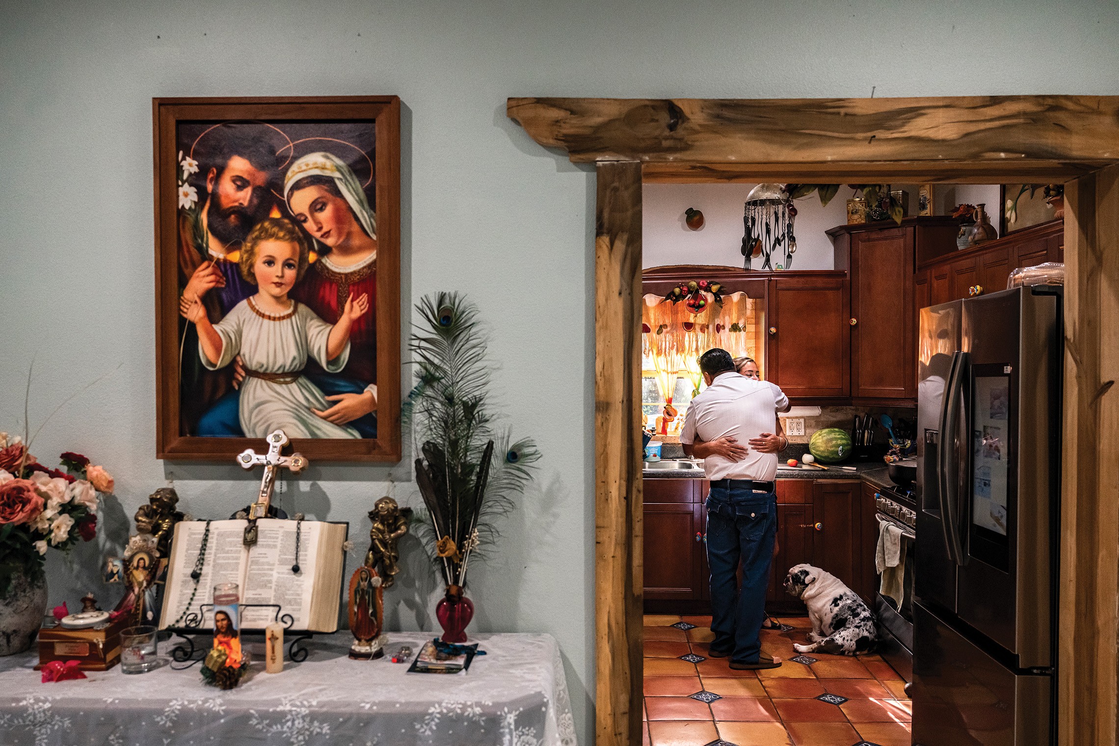 photo of room with painting of the holy family, side table with open bible and cross, and doorway looking into tiled kitchen, where two people embrace and a dog looks on