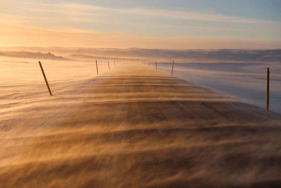 Wind blows snow across a road.