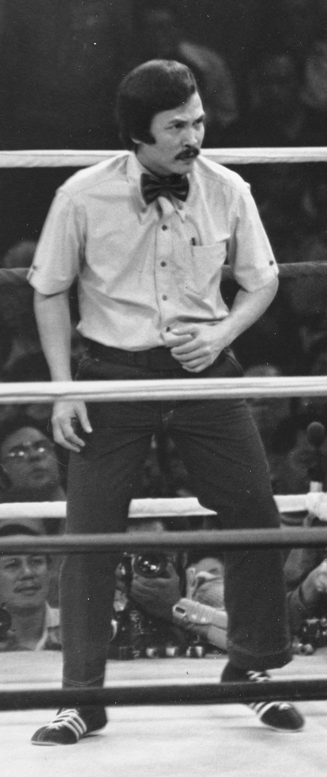 black-and-white photo of dark-haired man with dark mustache, wearing short-sleeve button-down shirt with bow tie and dark pants, inside boxing ring with crowd behind