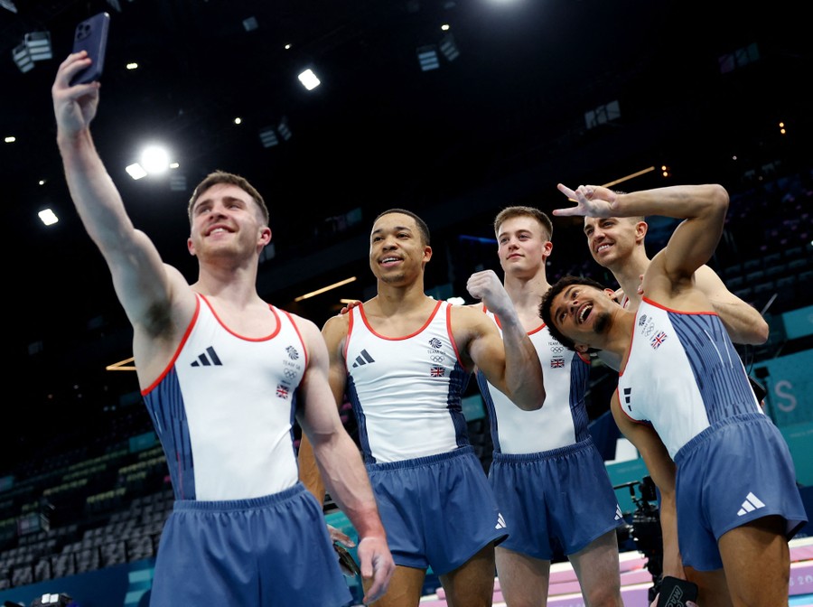 Five gymnasts pose for a selfie in an arena.