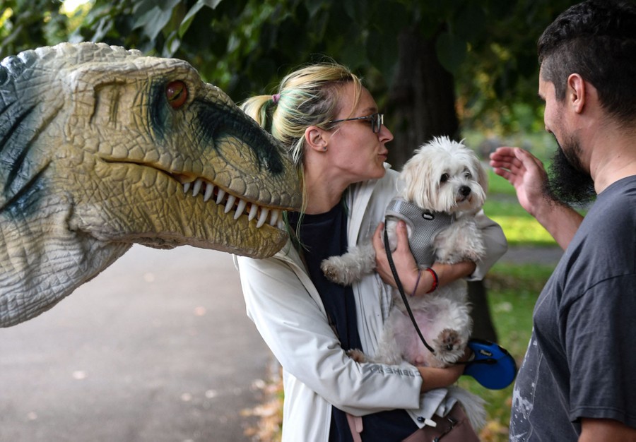 A person wearing a dinosaur costume stands beside a couple who are holding and comforting a small dog.