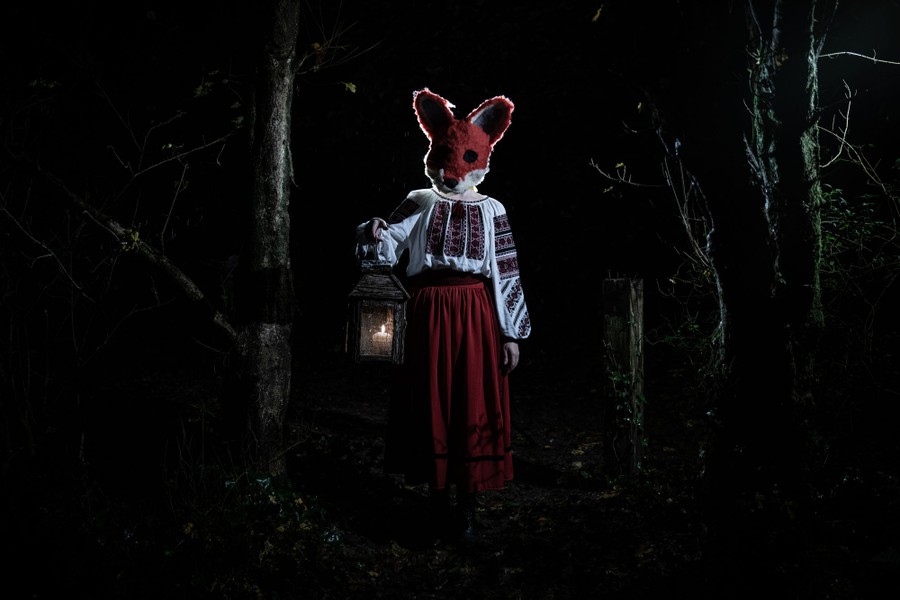 A person wearing a full-head fox mask carries a lantern in a wooded area.