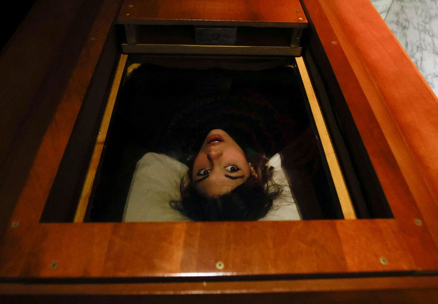 A person lies on their back in a coffin, captured through a hole in the top of the coffin, during an escape game.