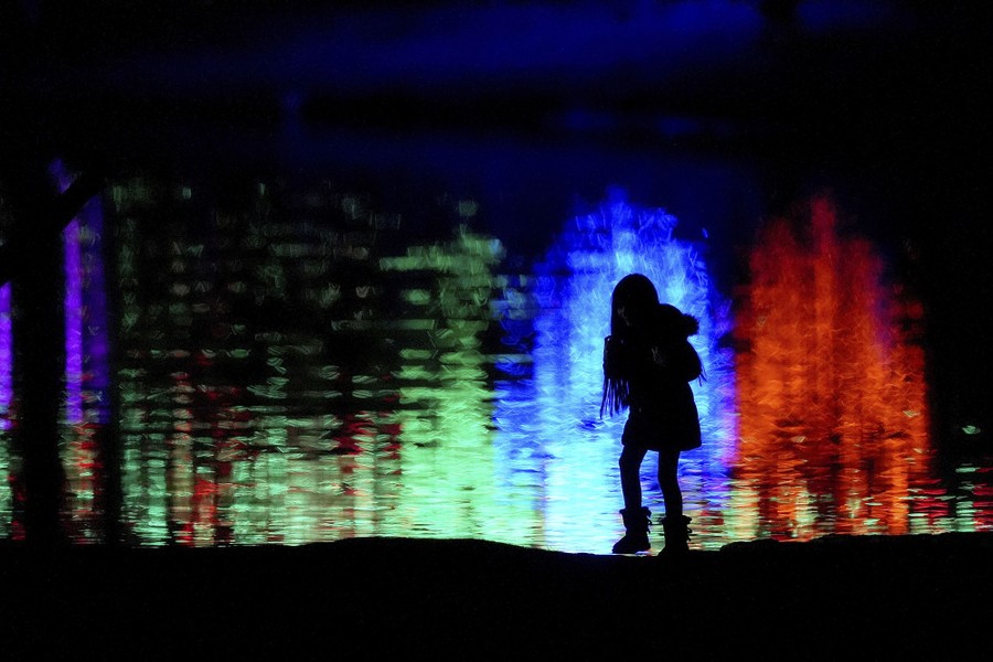 A child is silhouetted against Christmas lights reflected on a pond.
