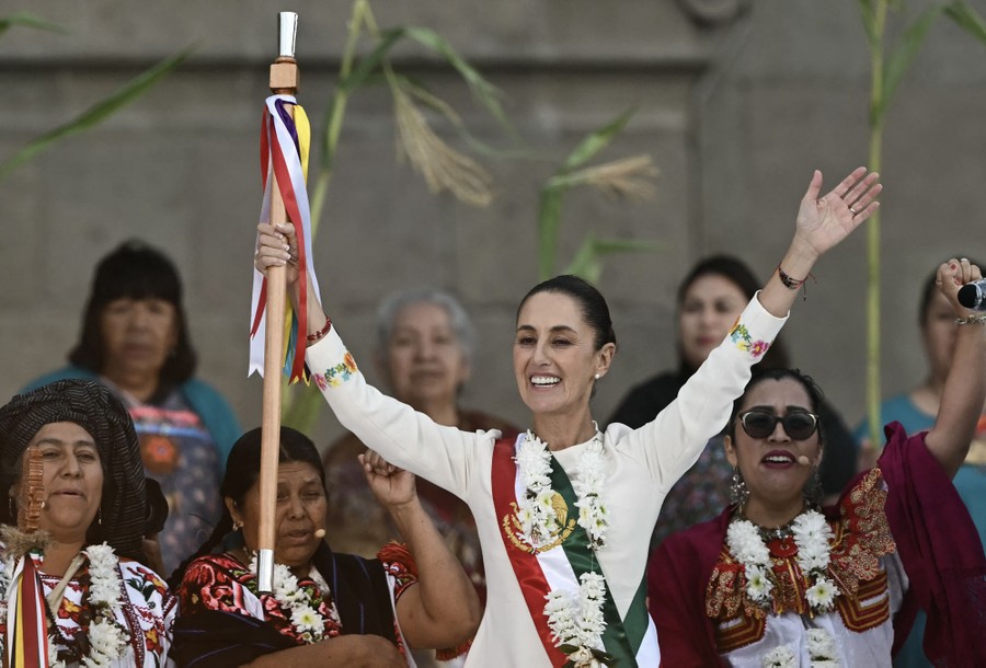A woman wearing a sash raises her arms, smiles, and holds up a ceremonial staff.