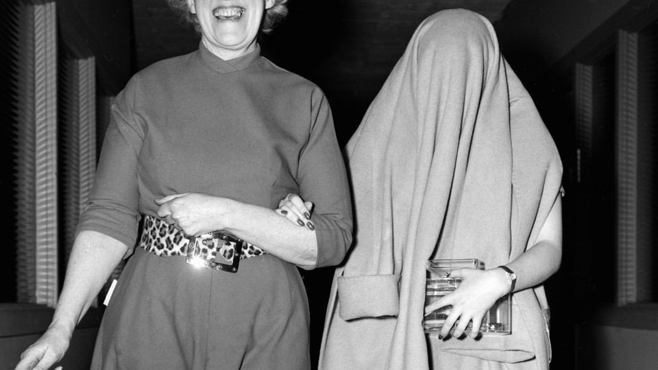 Black and white image of two women walking arm in arm, one with a big smile on her face, the other with a coat over her head, obscuring her face