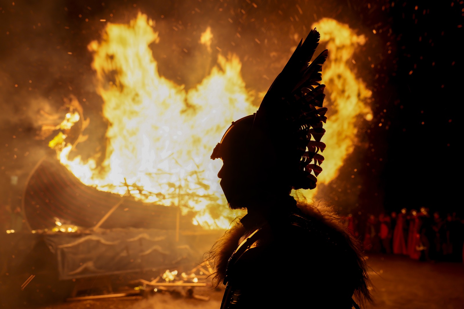 A person wearing a Viking costume with a winged helmet is seen in silhouette in front of a burning Viking ship.