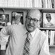 Black-and-white photo of the writer Roger Angell with baseball memorabilia and photos behind him on a bookshelf