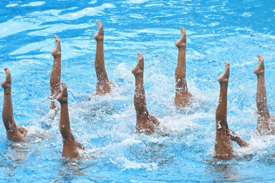 Eight artistic swimmers perform in a pool, in formation, each with one leg sticking out of the water.