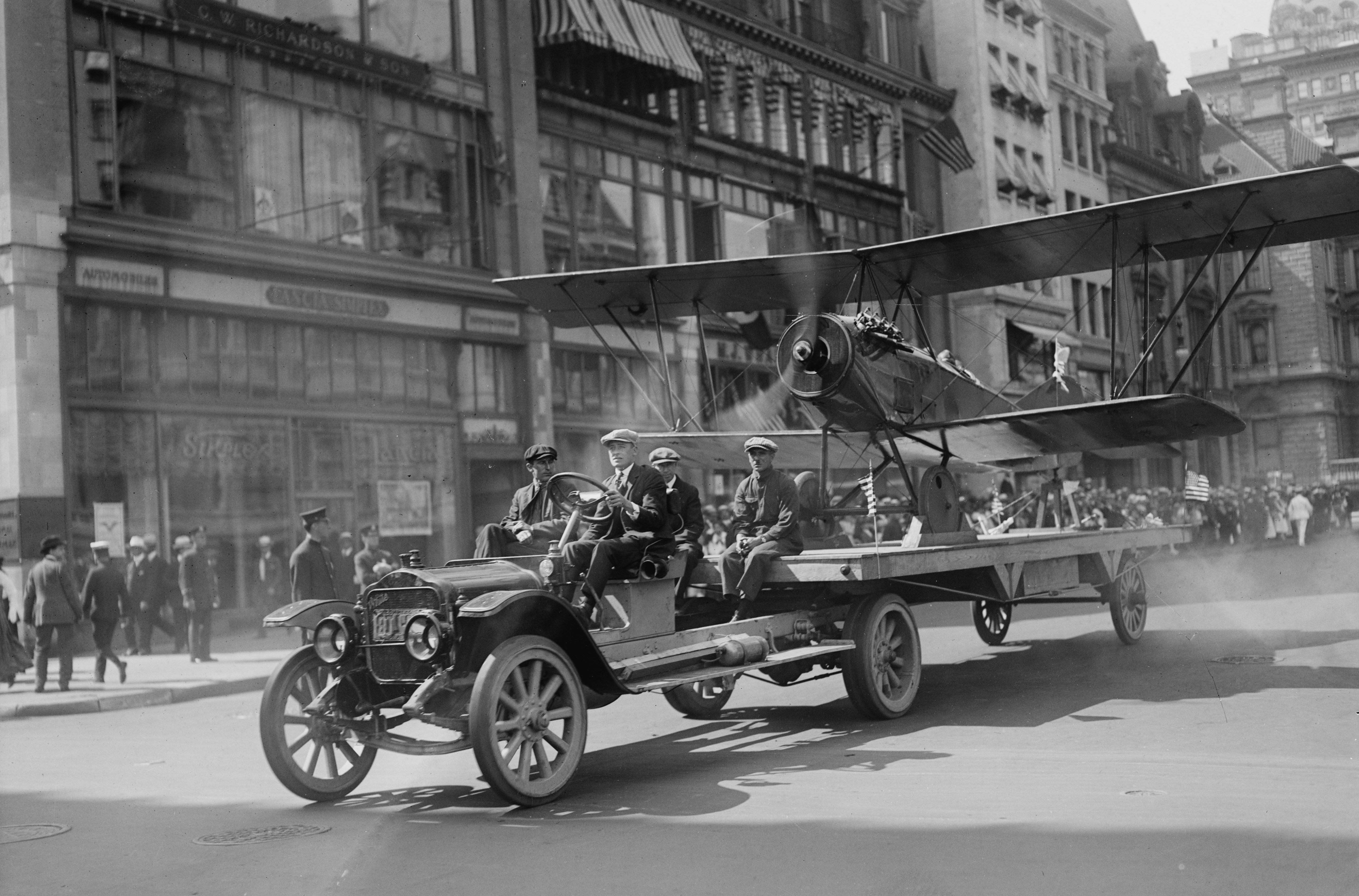 A truck tows a prop-motor biplane on a trailer in a parade in New York City in 1918. The propeller is spinning.