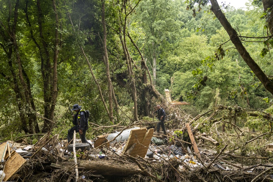 Two rescue workers walk through flood debris strewn across trees and bushes.