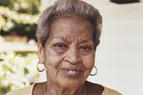 a portrait of Carmen Ayala outside the home where she and Adele lived. Carmen wears a yellow shirt and crucifix
