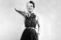 A black-and-white portrait of Claire McCardell, standing partially behind a frosted glass panel in a black dress, her left hand tucked into a side pocket.
