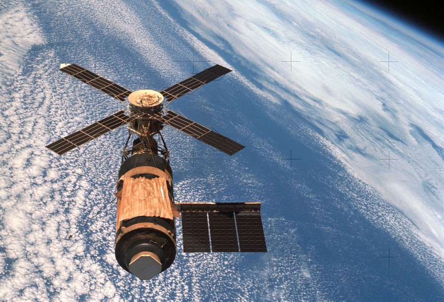 A spacecraft, shaped like a long cylinder with several outstretched solar panels, is seen above clouds, orbiting the Earth.