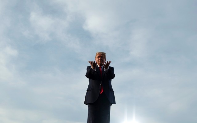 Donald Trump stands against a blue sky, gesturing toward the viewer.