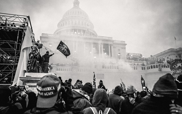 A photo of a crowd with a MAGA hat in the foreground and people standing on scaffolding with a Second Amendment flag and Trump banner in front of the smoke-obscured U.S. Capitol