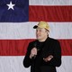 Elon Musk shrugs wearing a MAGA hat in front of an American flag