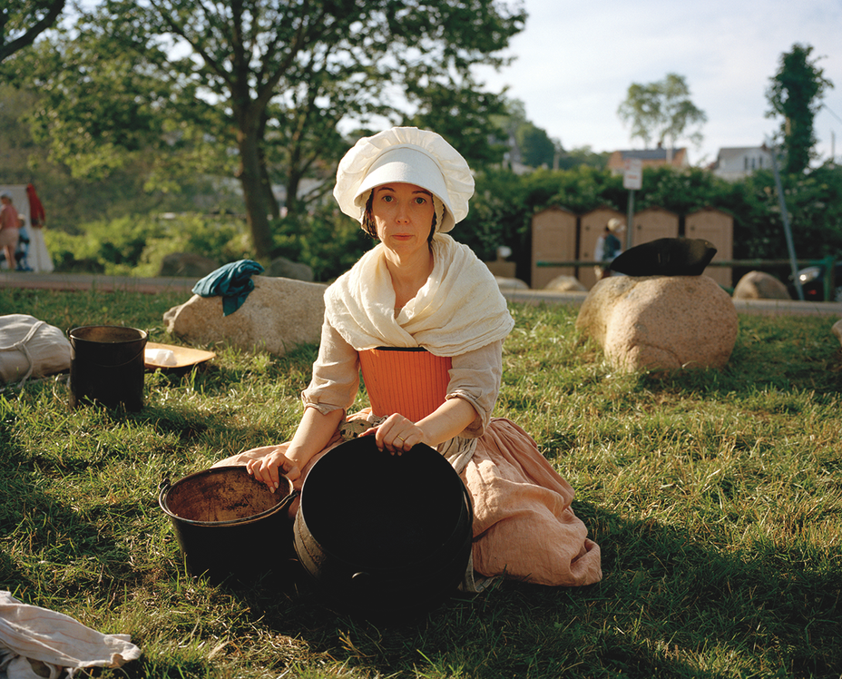 photo of a woman in white colonial bonnet, white shawl, and pink dress kneeling in grass holding two baskets while doing the washing at a reenactment of Bunker Hill in Gloucester, MA