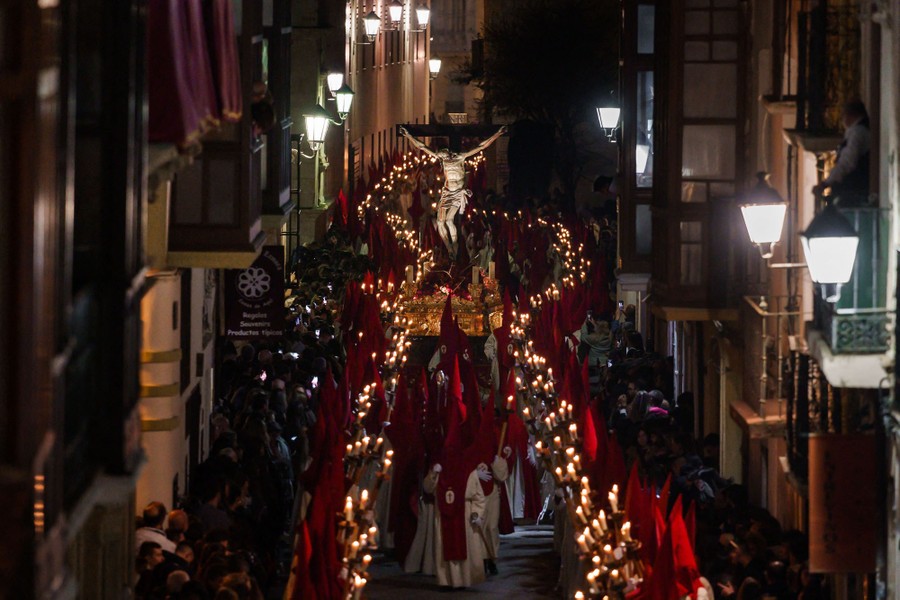 A line of penitents carrying candles marches down a street carrying a statue of the crucified Jesus Christ.