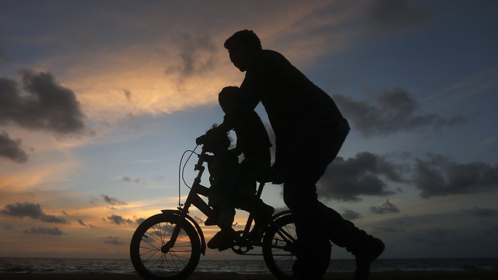 A man guides a child riding a two-wheel bike. It's sunset, and the figures appear as silhouettes.